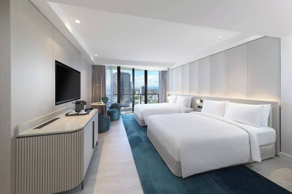 A modern hotel room with a large bed, a desk with a flat-screen TV, and a view of the cityscape.