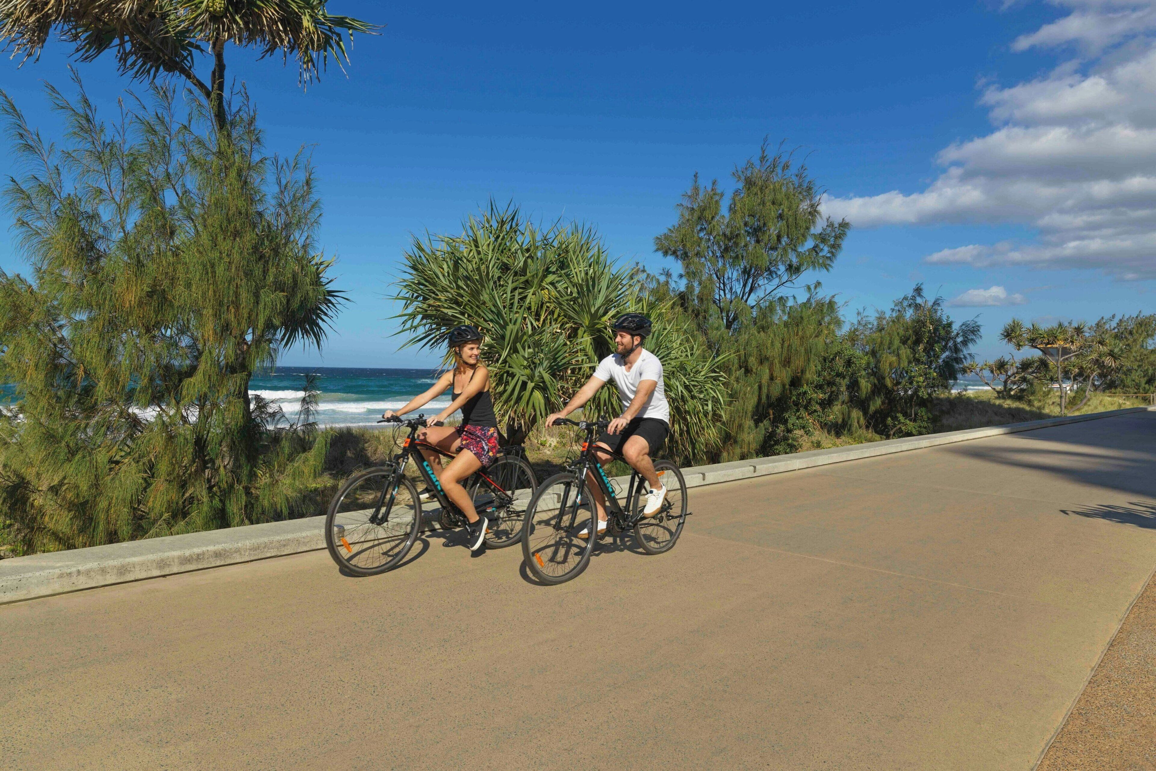 Dos personas andan en bicicleta por un camino pavimentado con vista al océano y a los árboles.