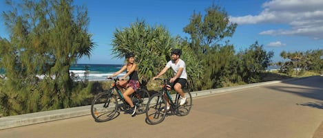 Dos personas andan en bicicleta por un camino pavimentado con vista al océano y a los árboles.