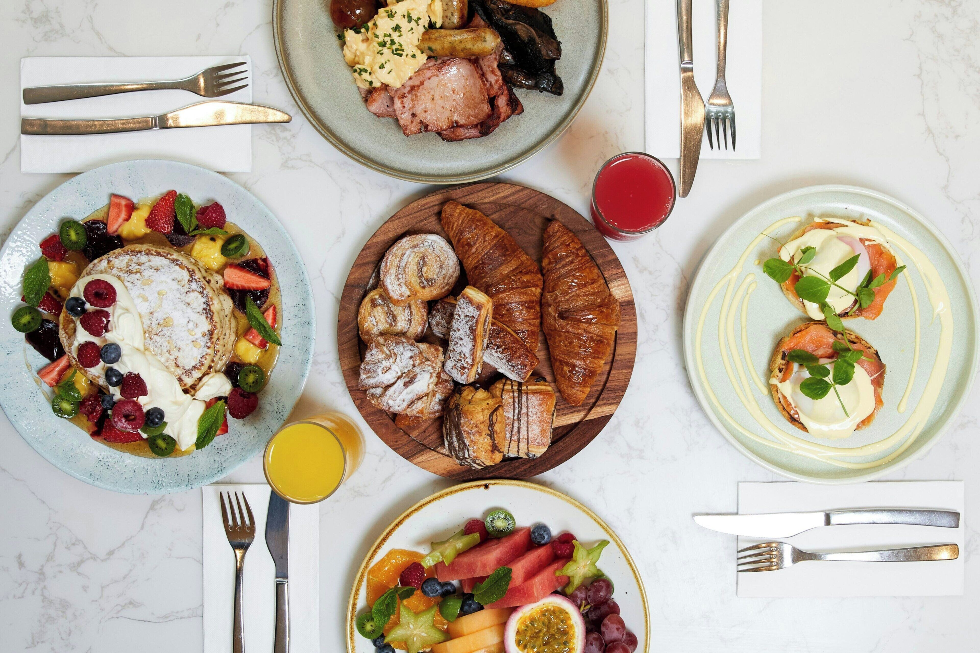 En frukostuppdukning med olika rätter, inklusive pannkakor, croissanter och frukt.