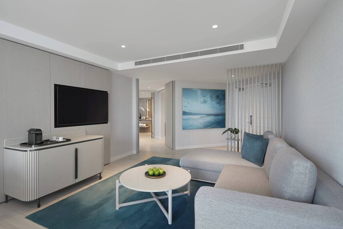 A modern living room with a grey sofa, a round coffee table with apples, a flat-screen TV, and a painting on the wall.