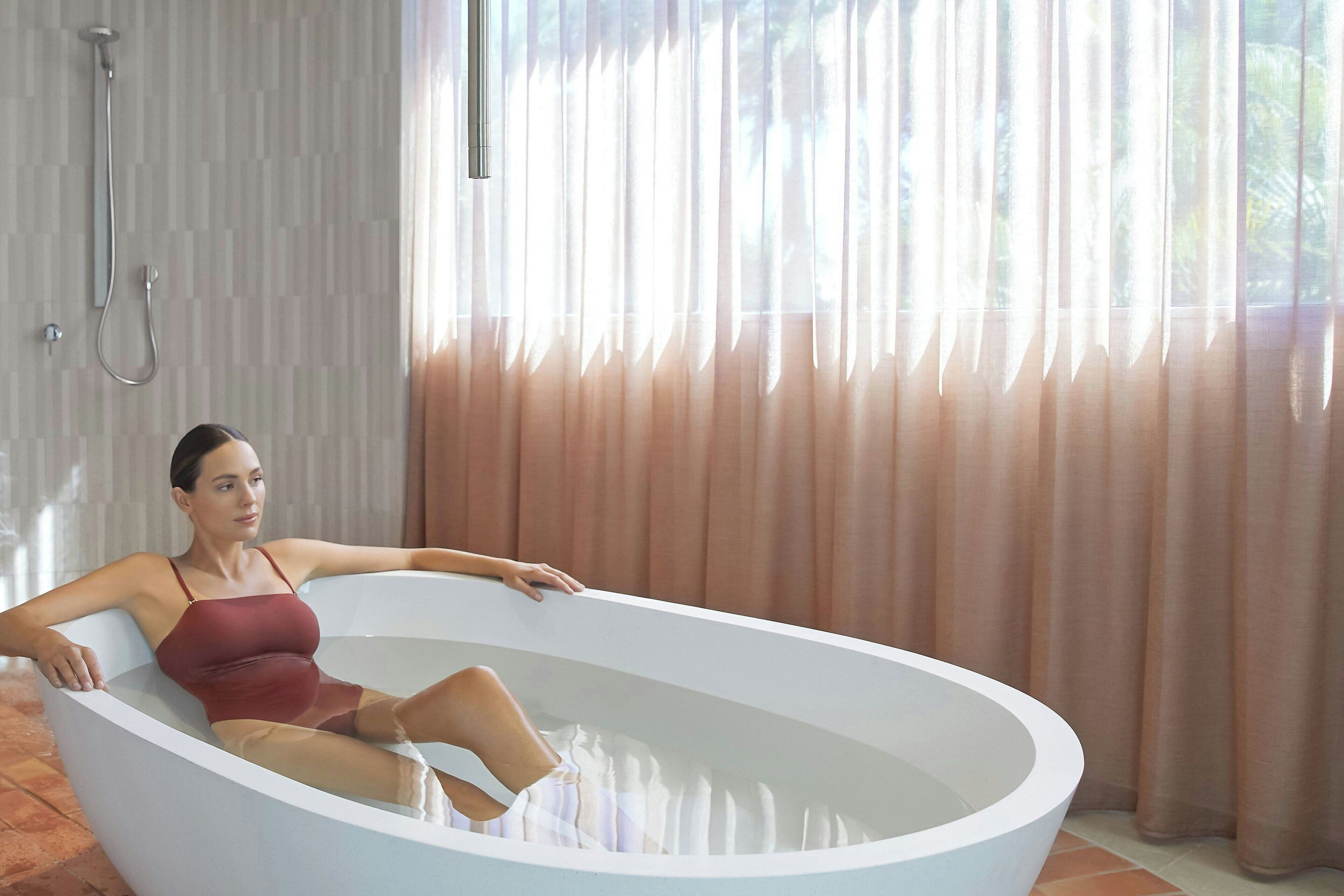 A woman in a bathtub in a modern bathroom with a showerhead and curtains.