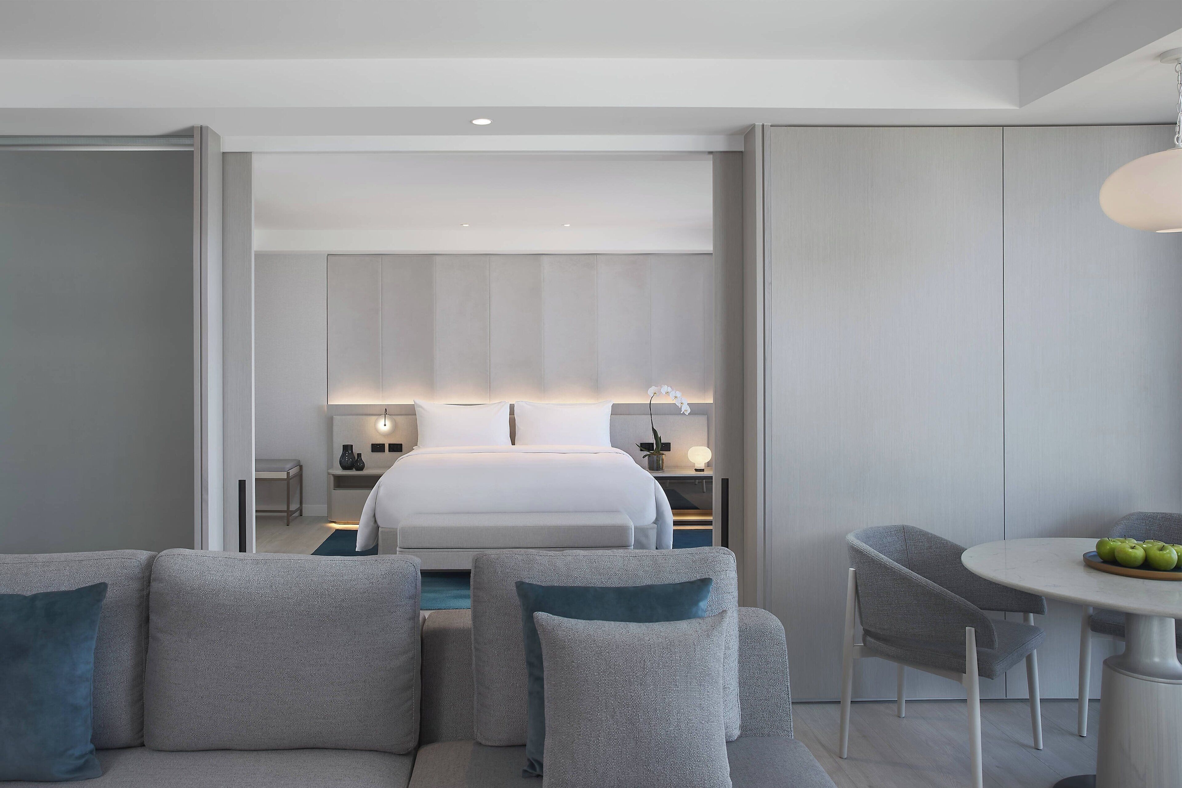 A modern hotel room with a large bed, a seating area with sofas, a dining table with apples, and a small kitchen area.