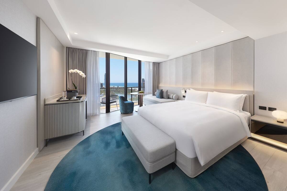 A modern hotel room with a large bed, a seating area with a chair, a small table, and a view of the city and water.