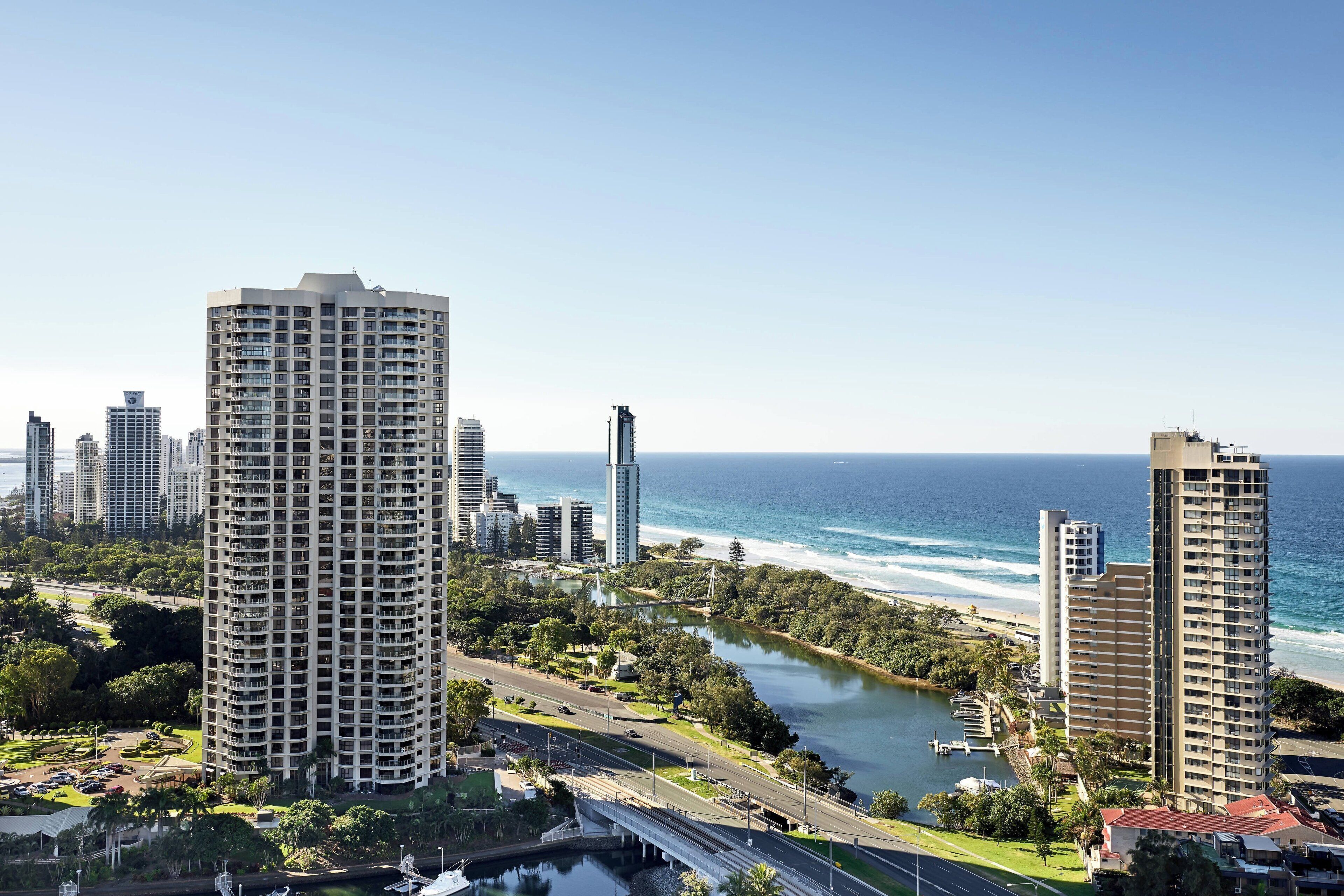 A high-rise hotel with multiple floors, a view of the ocean, and a park area with a bridge.