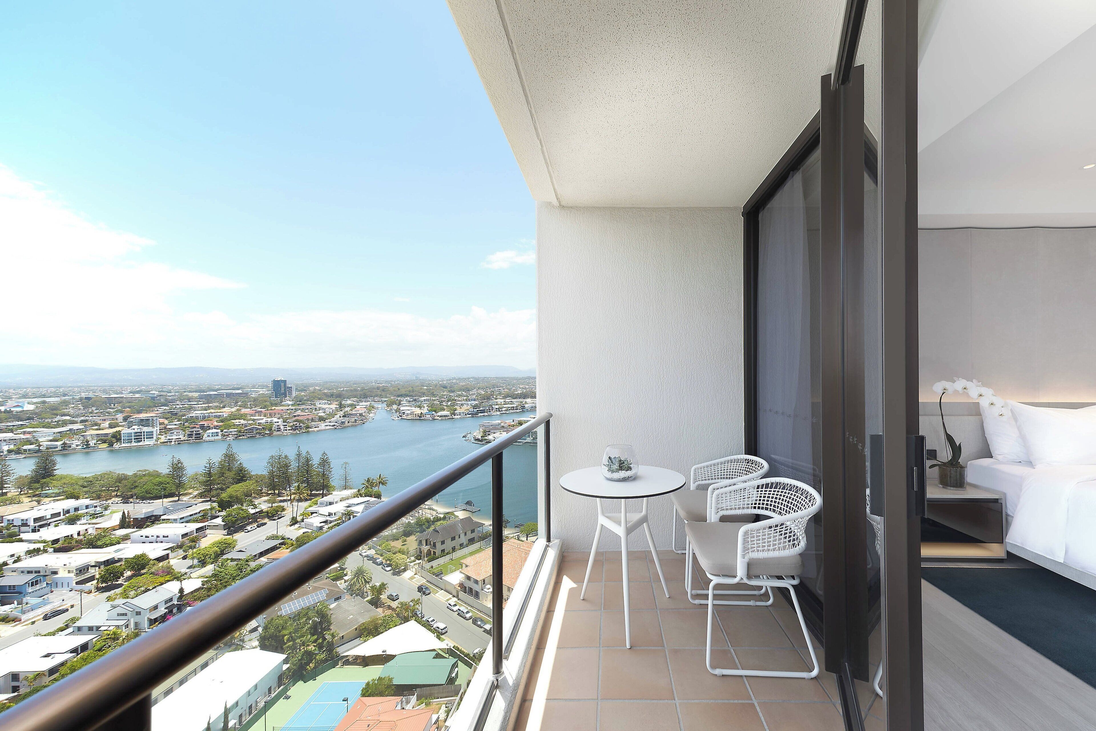 A balcony with a view of a lake and cityscape, a small table with chairs, and a bed with white bedding.