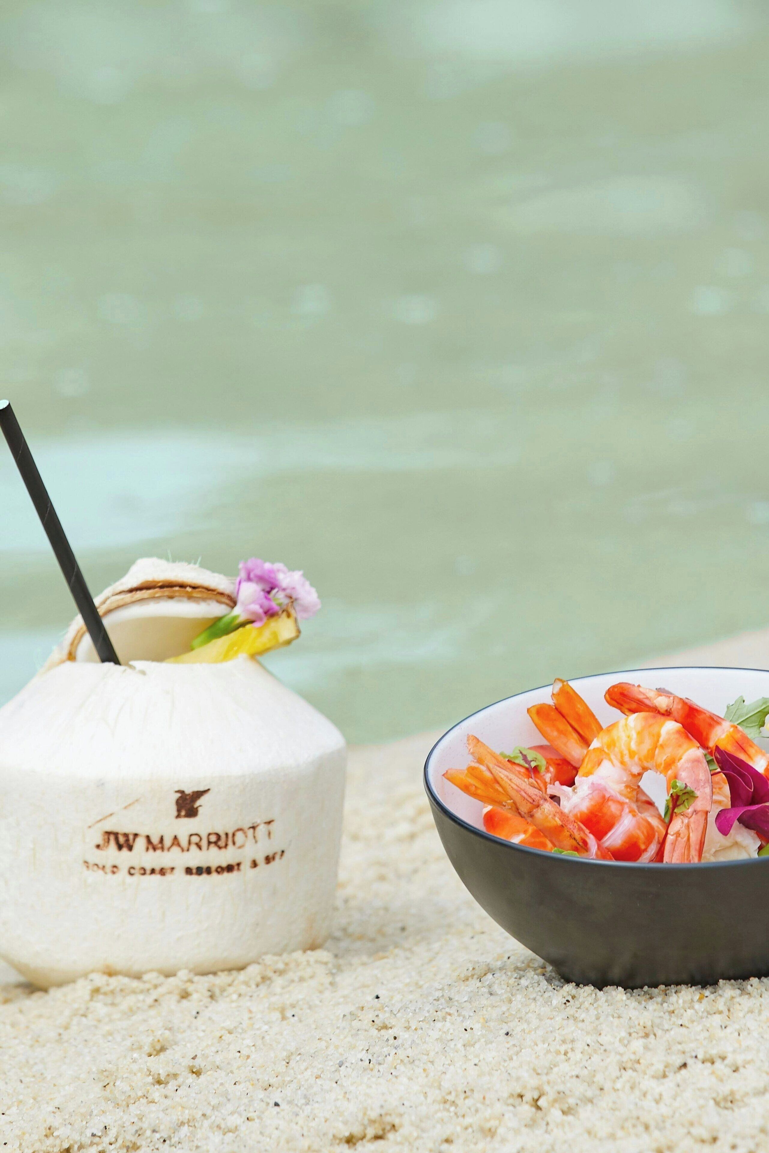 A coconut drink with a straw and a flower, and a bowl of shrimp salad on a sandy surface.