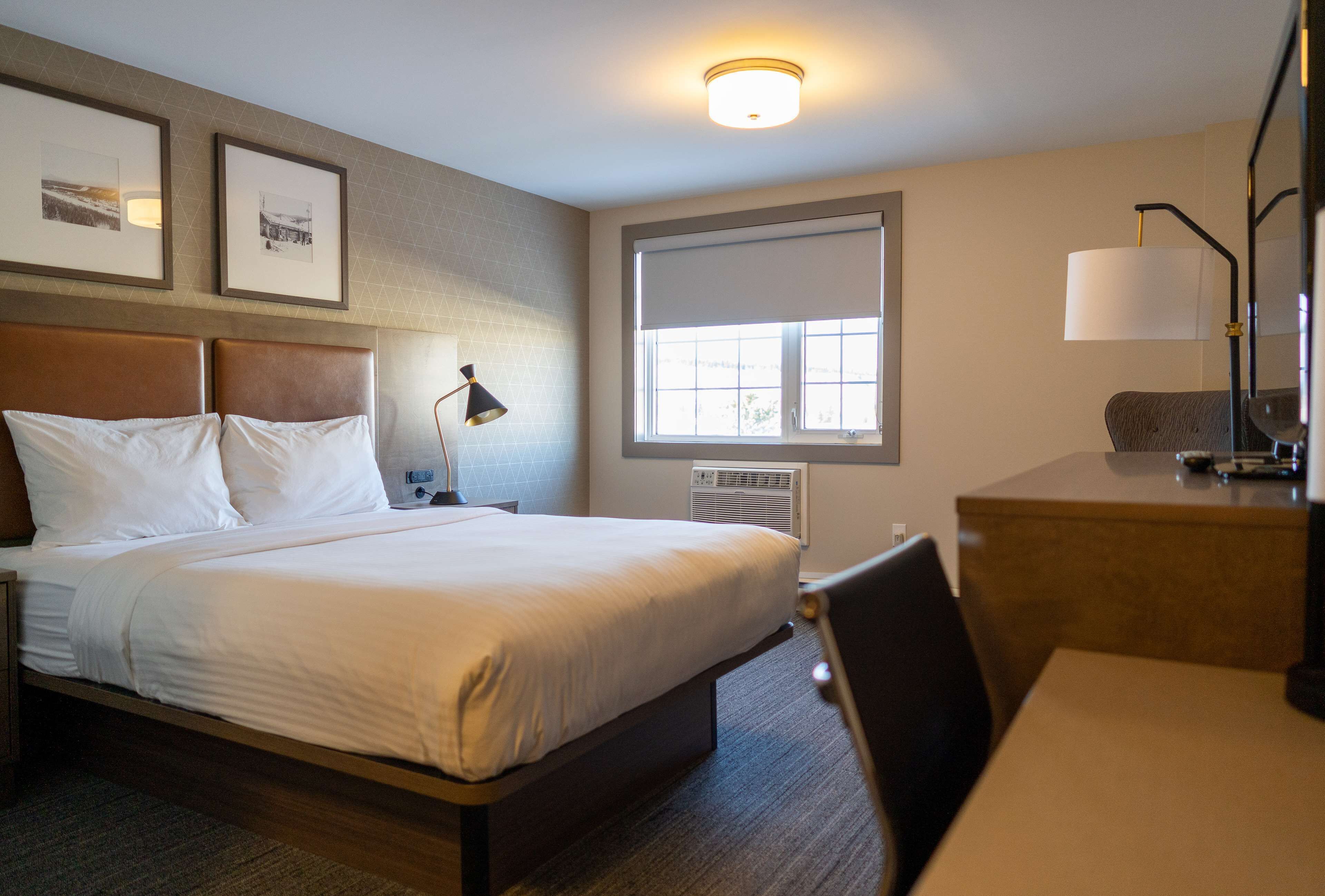 Une chambre d’hôtel comprenant un lit, une table de chevet, une lampe, un bureau, une chaise et une fenêtre avec des stores.