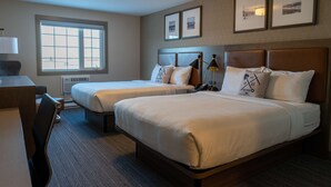 Une chambre d’hôtel avec deux lits, un bureau et une chaise. Les murs sont ornés de tableaux encadrés, il y a une fenêtre et un luminaire au plafond.