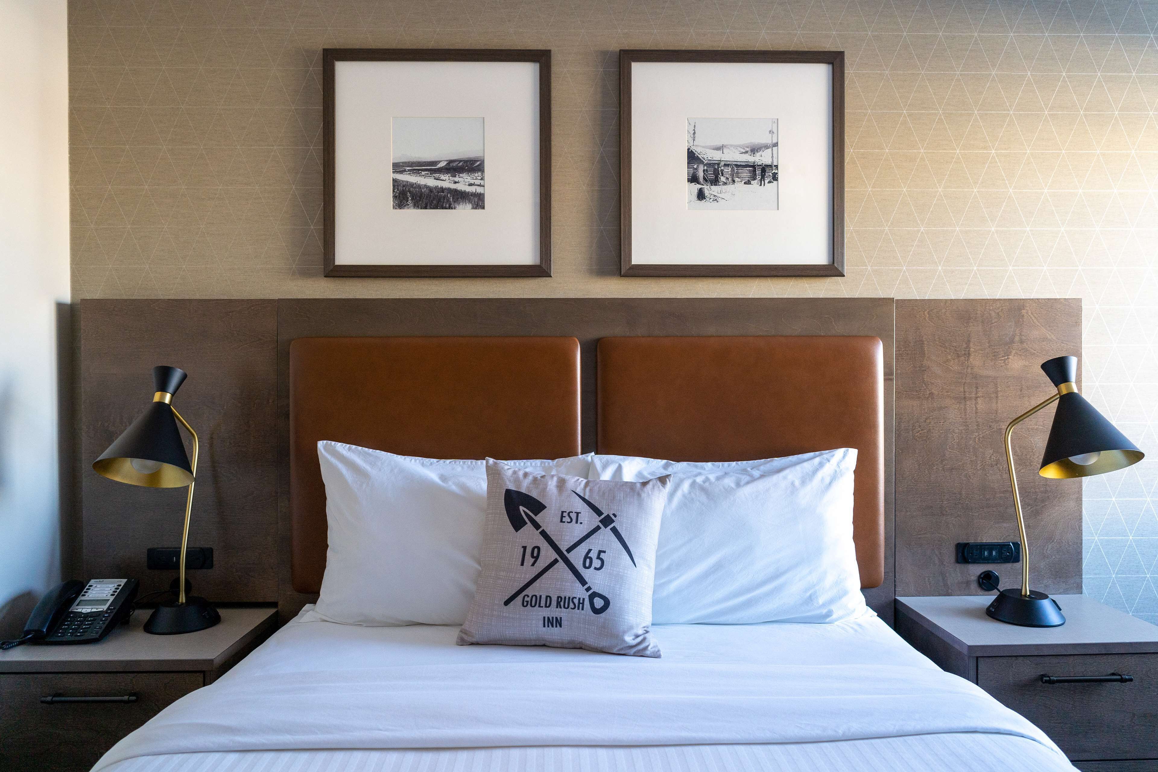 A neatly made bed with a pillow featuring a logo, two framed pictures on the wall, and bedside lamps on nightstands.