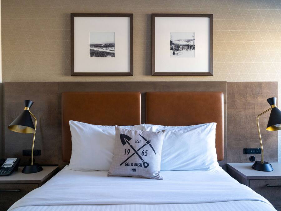 Een net opgemaakt bed met een kussen met een logo, twee ingelijste foto’s aan de muur en nachtlampjes op nachtkastjes.