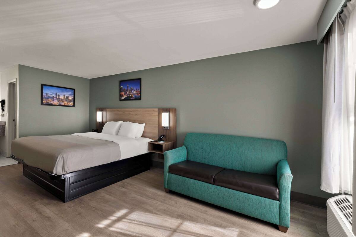 A hotel room with a bed, a teal sofa, two framed pictures on the wall, and a window with curtains.