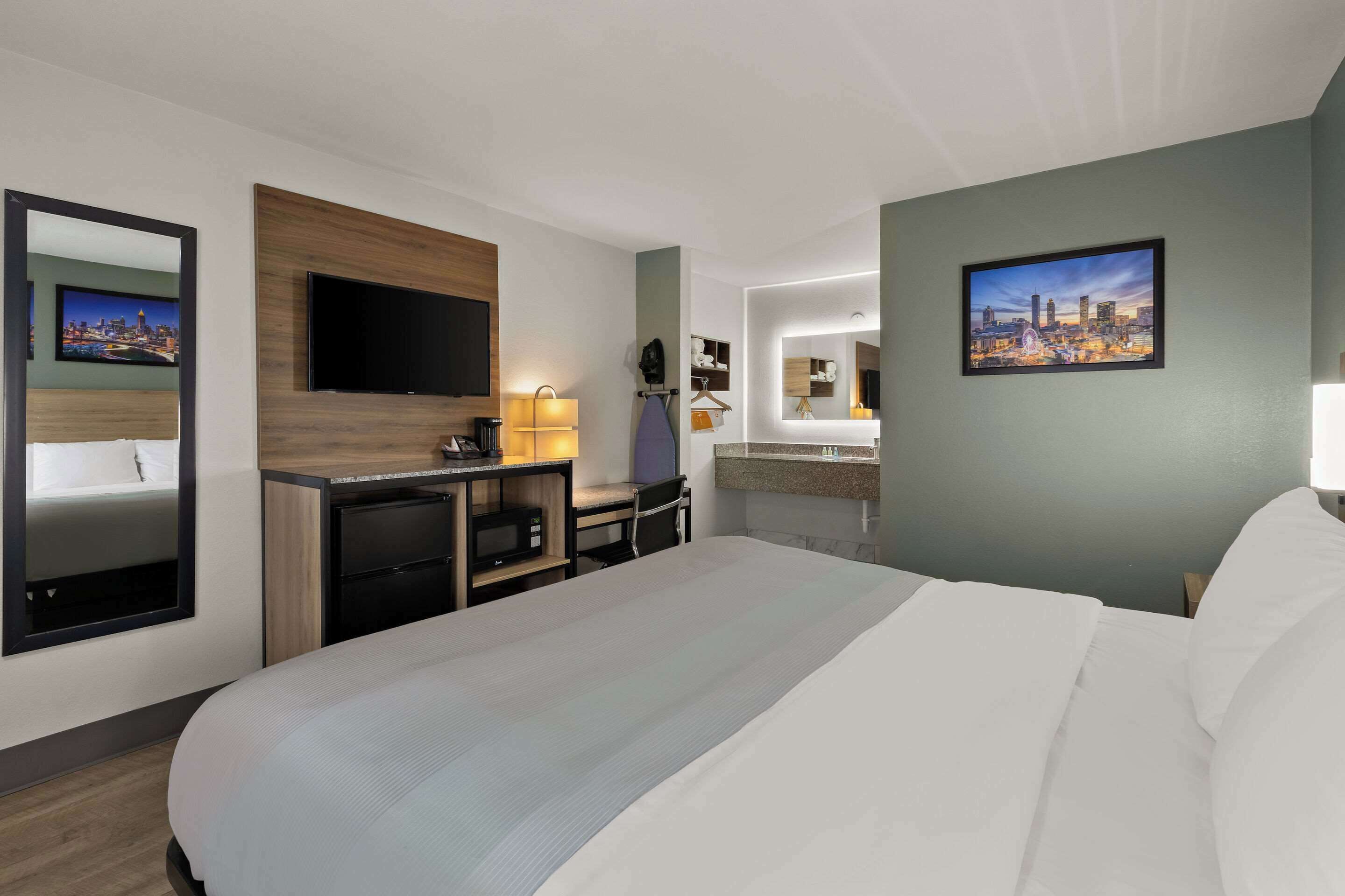 Ein Hotelzimmer mit einem großen Bett, einem Schreibtisch mit Computer, einem flachbildfernseher an der Wand und einem Badezimmer mit Spiegel und Waschbecken.