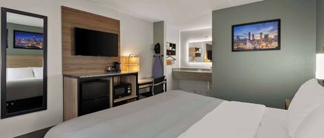 Ein Hotelzimmer mit einem großen Bett, einem Schreibtisch mit Computer, einem flachbildfernseher an der Wand und einem Badezimmer mit Spiegel und Waschbecken.