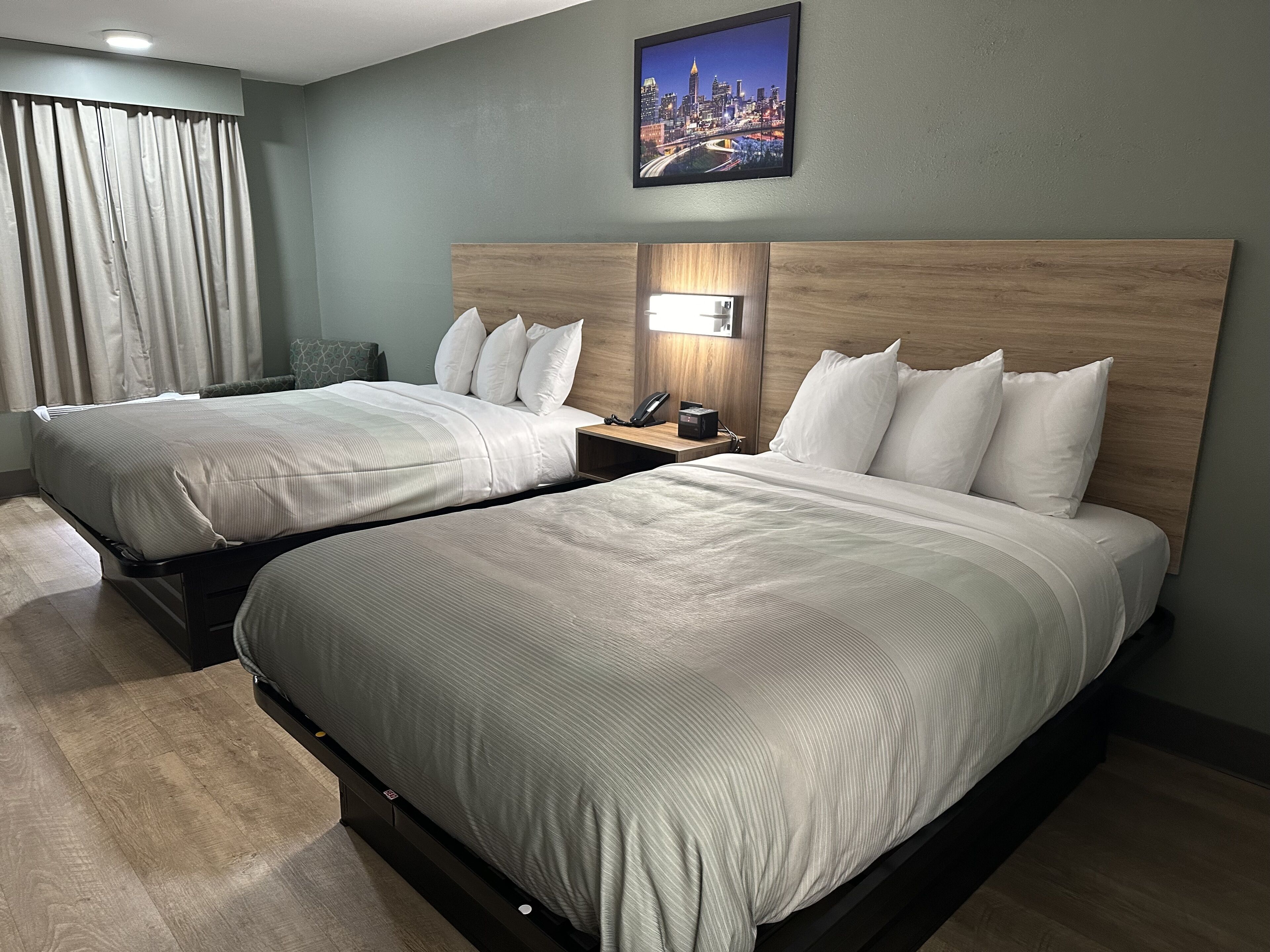 A hotel room with two beds, a cityscape painting, a nightstand with a lamp, and a view of the city through the window.