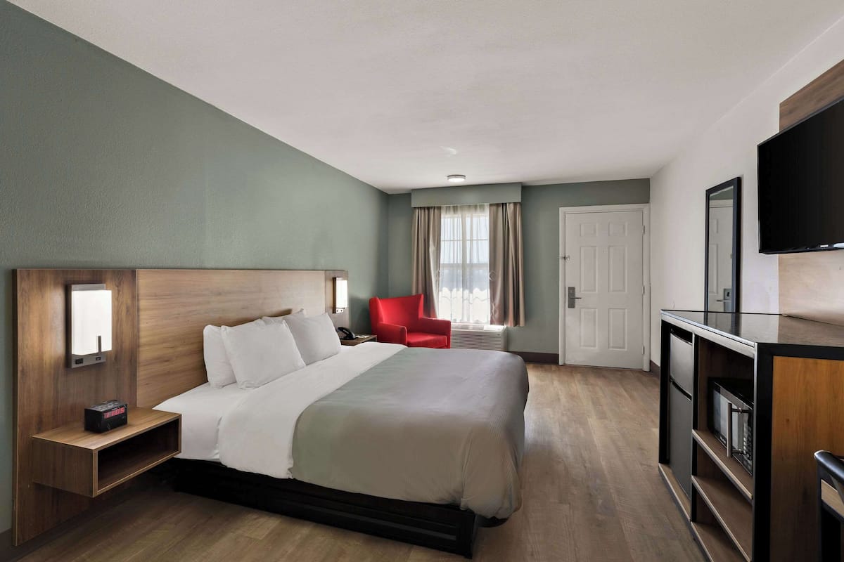 A hotel room with a large bed, a red chair, a TV, and a desk with a lamp.