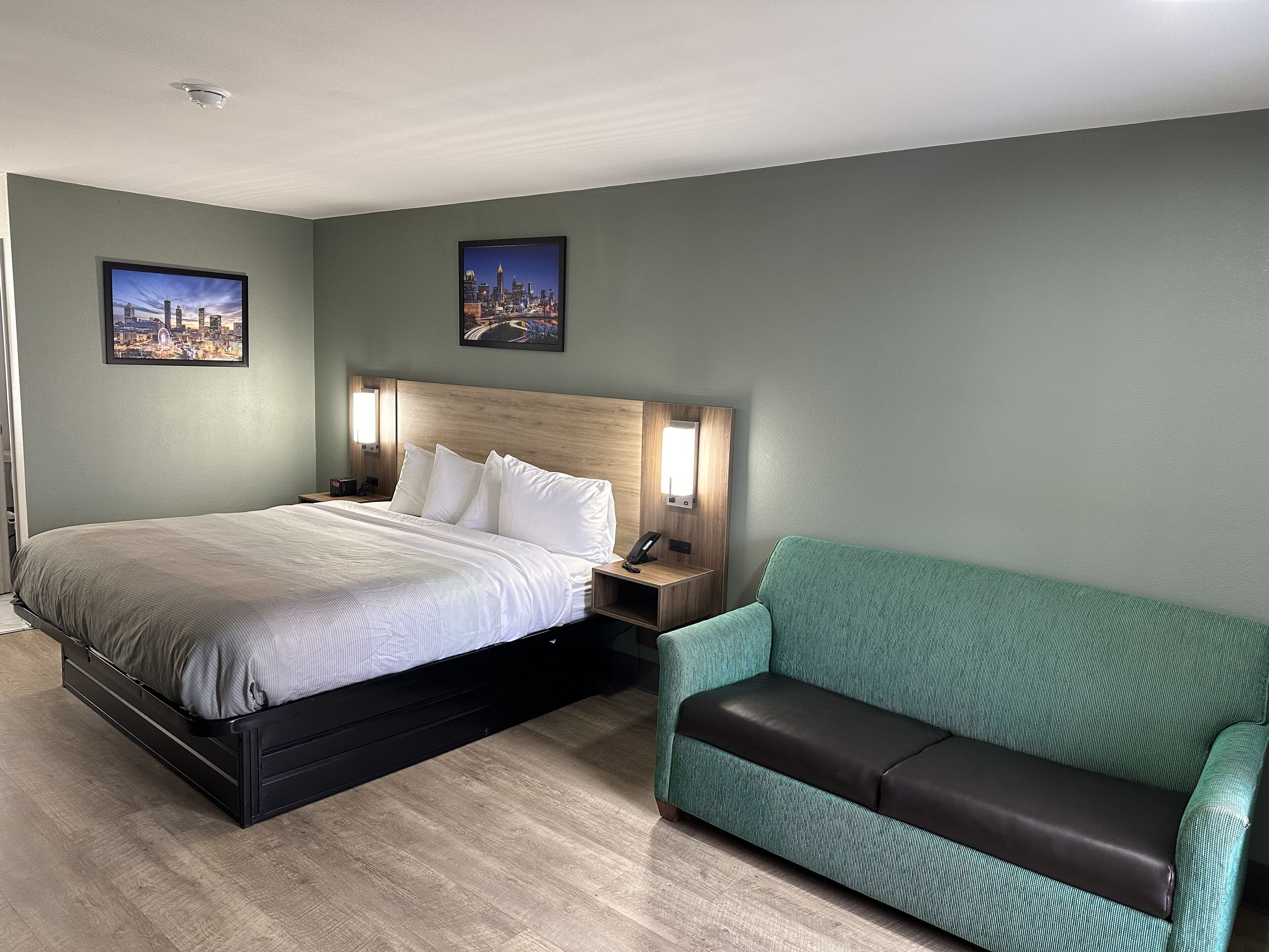 Une chambre d’hôtel avec un lit, une table de chevet, un canapé vert et deux tableaux représentant des paysages urbains accrochés au mur.