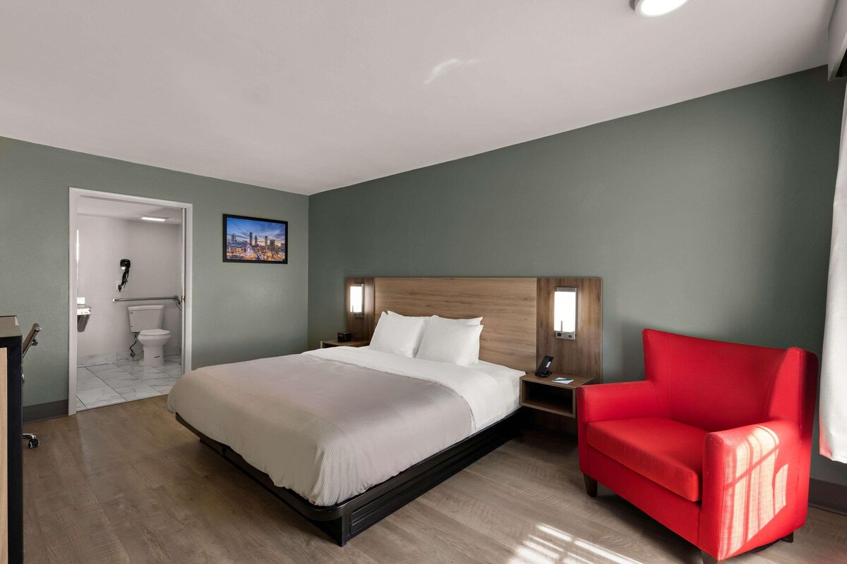 Habitación de hotel con cama, sillón rojo, mesita de noche, televisor montado en la pared y baño visible a través de una puerta abierta.