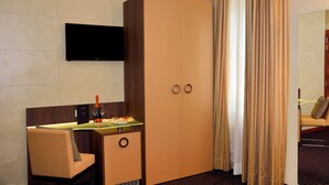 A hotel room with a large wardrobe, a desk with a bottle and glasses, a television mounted on the wall, and a bed with a headboard. - LH Hotel Sirio Venice (Mestre)