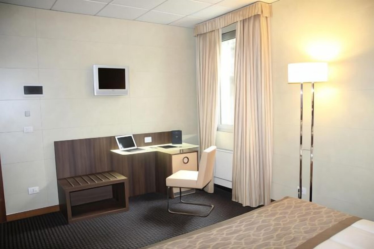 A hotel room with a bed, a desk with a laptop, a chair, a bedside table, and a lamp.