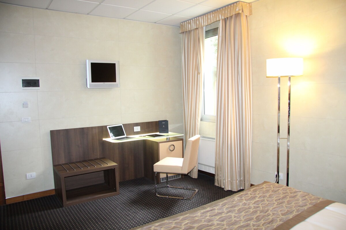 A hotel room with a bed, a desk with a laptop, a chair, a bench, a TV, and a window with curtains.
