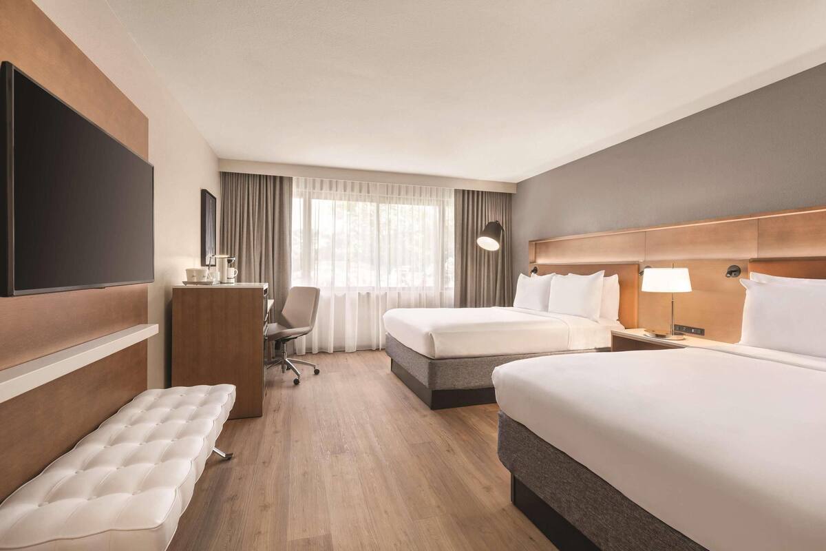 A hotel room with two beds, a desk, a chair, a TV, and a window with curtains.