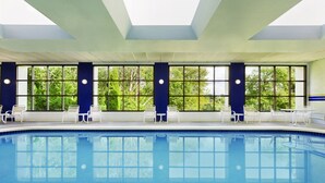 A swimming pool with clear blue water, surrounded by white lounge chairs and tables, large windows with a view of greenery, and a high ceiling with skylights. - Radisson Hotel Seattle Airport (SeaTac)