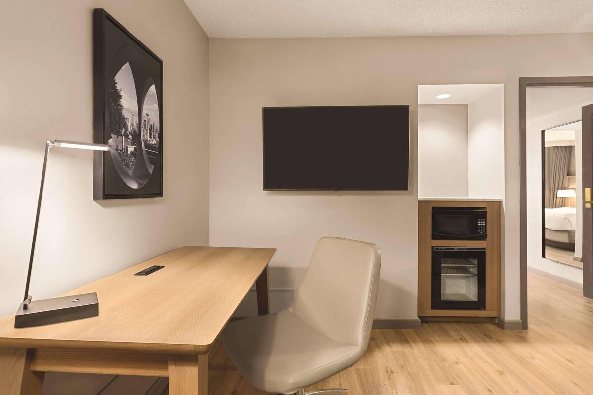 A room with a wooden desk, a chair, a mounted flat-screen TV, a microwave, and an oven built into a cabinet.
