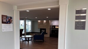 A spacious living area with a stone fireplace, a dining table, and blue chairs. There are signs indicating room numbers and amenities like a meeting room and ice machine. - Wingate by Wyndham Dayton North (Dayton)