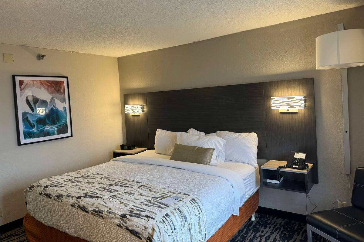 Ein Hotelzimmer mit einem großen Bett, einem Nachttisch mit Lampe, einem Telefon und einer Wandleuchte.