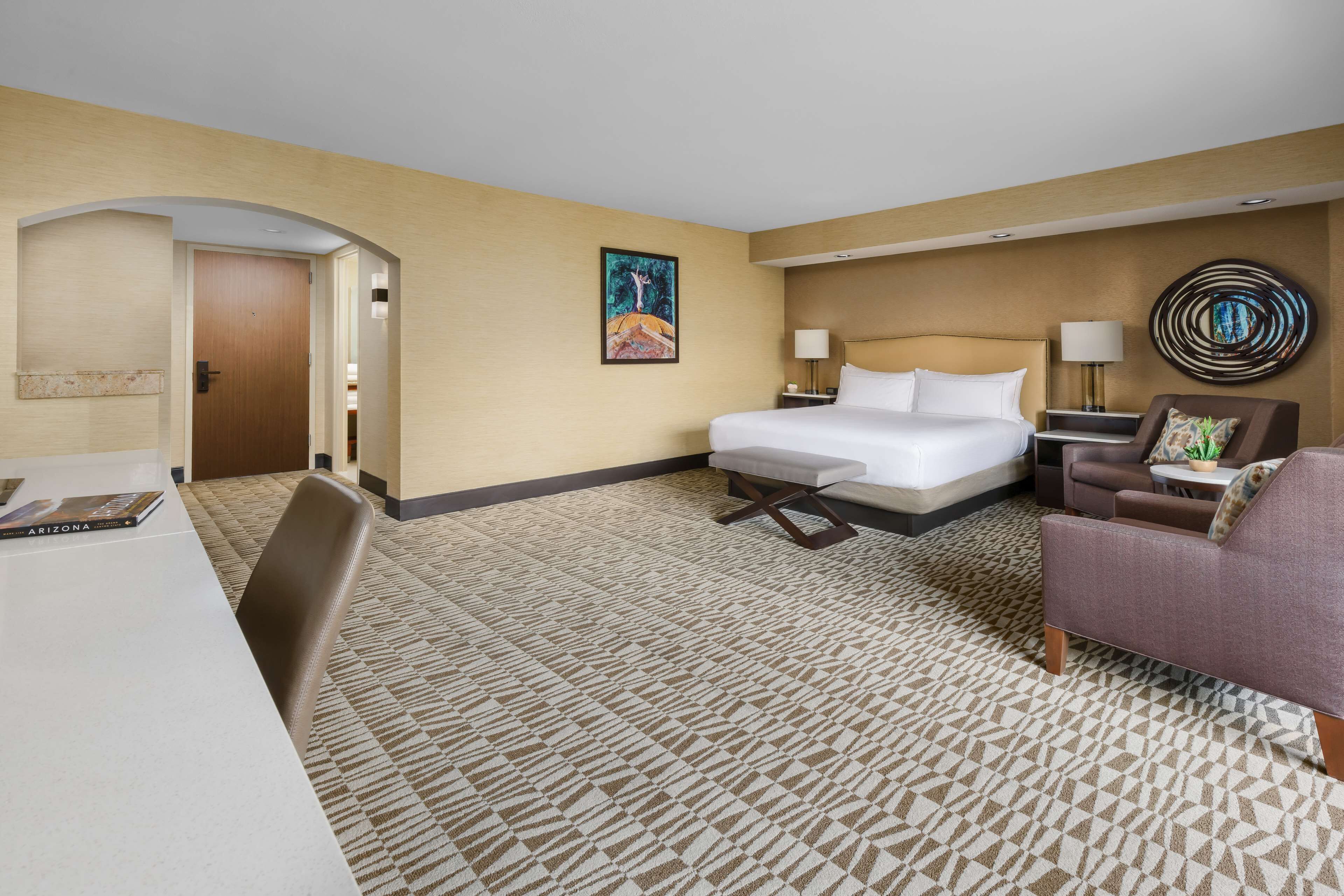 Una habitación de hotel con una cama, un escritorio, una silla, un sofá, una lámpara y un cuadro en la pared.