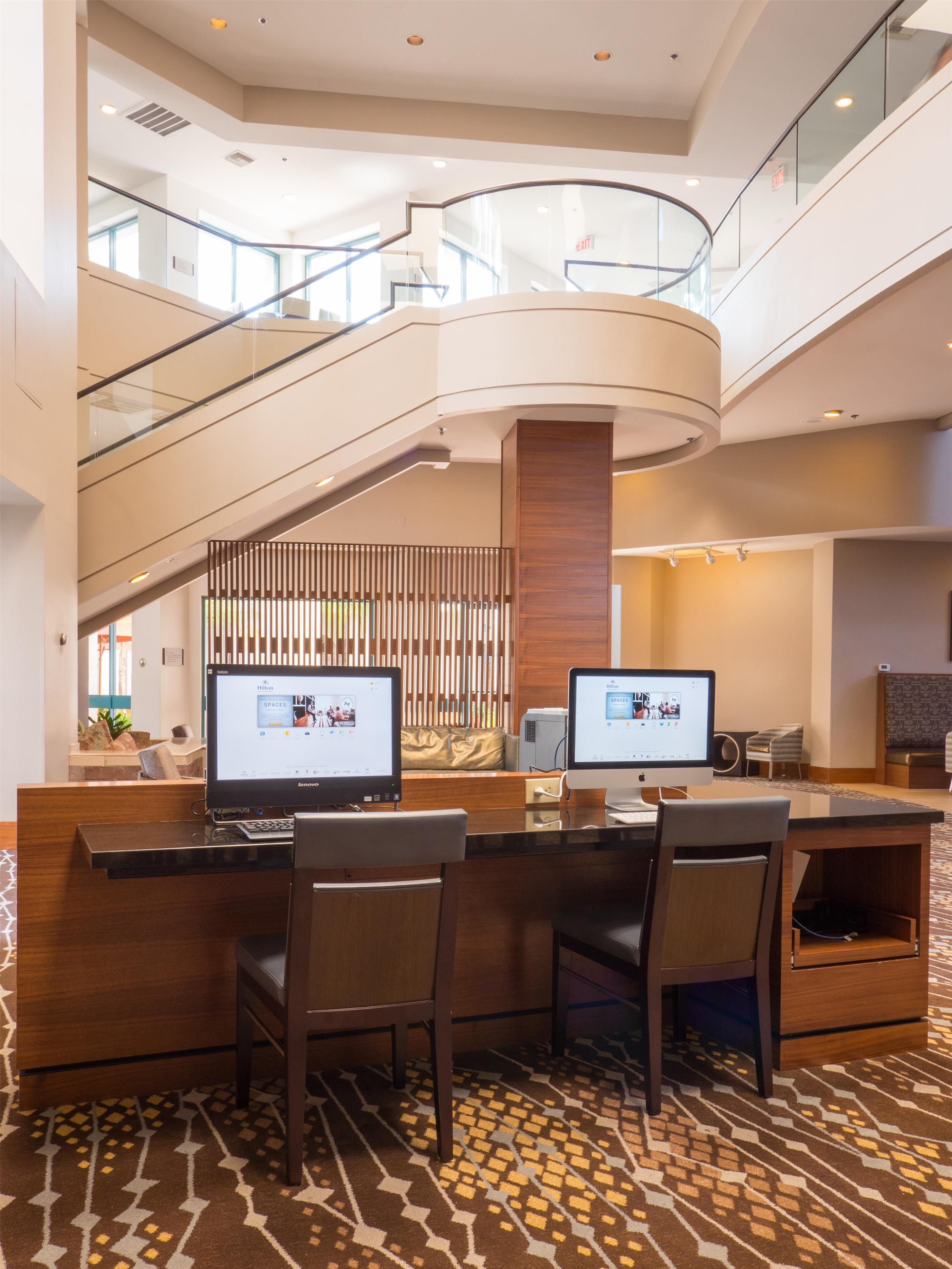 A hotel lobby with a reception desk, two computer monitors, a staircase, and a patterned carpet.