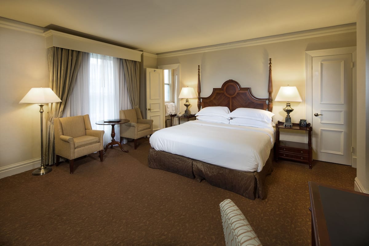 Ein Hotelzimmer mit einem großen Bett, zwei Nachttischen mit Lampen, einem Sessel, einem kleinen runden Tisch und einem Schreibtisch.