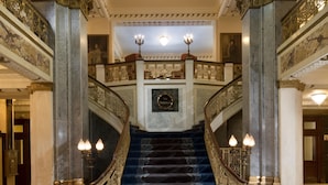 Un magnifique escalier orné de balustrades travaillées et doté d’un sol en marbre, menant à un hall spacieux à hauts plafonds, agrémenté de moulures décoratives et d’un lustre.