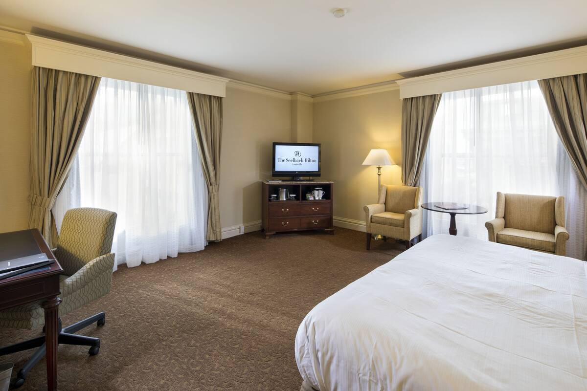 Ein Hotelzimmer mit einem großen Bett, einem Schreibtisch mit einem Laptop, einem Fernseher, einem Sessel, einer Lampe und einem kleinen Tisch.