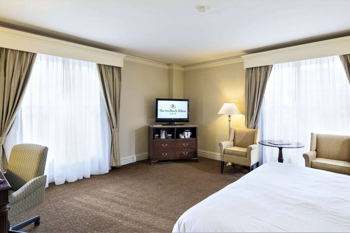 Ein Hotelzimmer mit einem Bett, zwei Sesseln, einem Schreibtisch mit einem Laptop, einem Fernseher und einer Lampe.