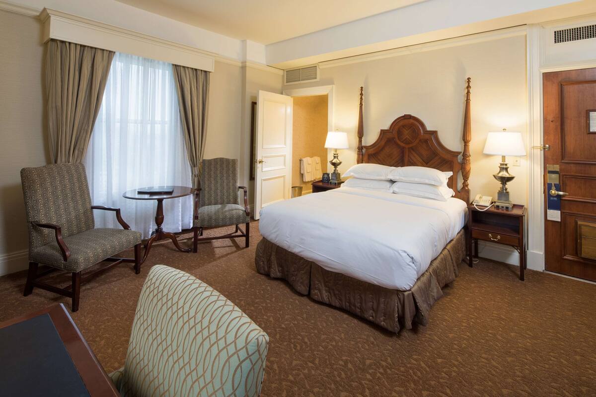 Ein Hotelzimmer mit einem großen Bett, zwei Sesseln, einem kleinen Tisch und einem Nachttisch mit Lampen.