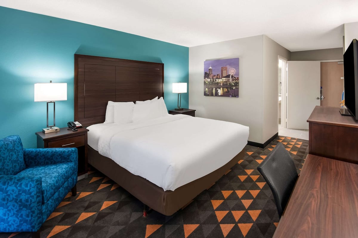 A hotel room with a large bed, two bedside tables, a desk, a chair, a TV, and a cityscape painting on the wall.