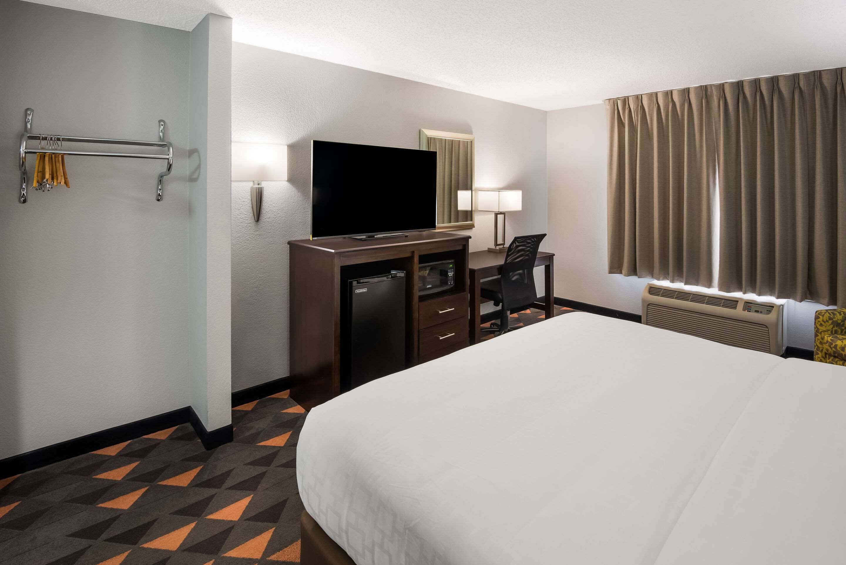A hotel room with a large bed, a desk with a flat-screen TV, a minibar, a seating area with a chair, and a window with curtains.