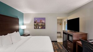 A hotel room with a bed, a desk with a TV, a lamp, and a cityscape painting on the wall. - Clarion Pointe Indianapolis Northeast (Indianapolis)