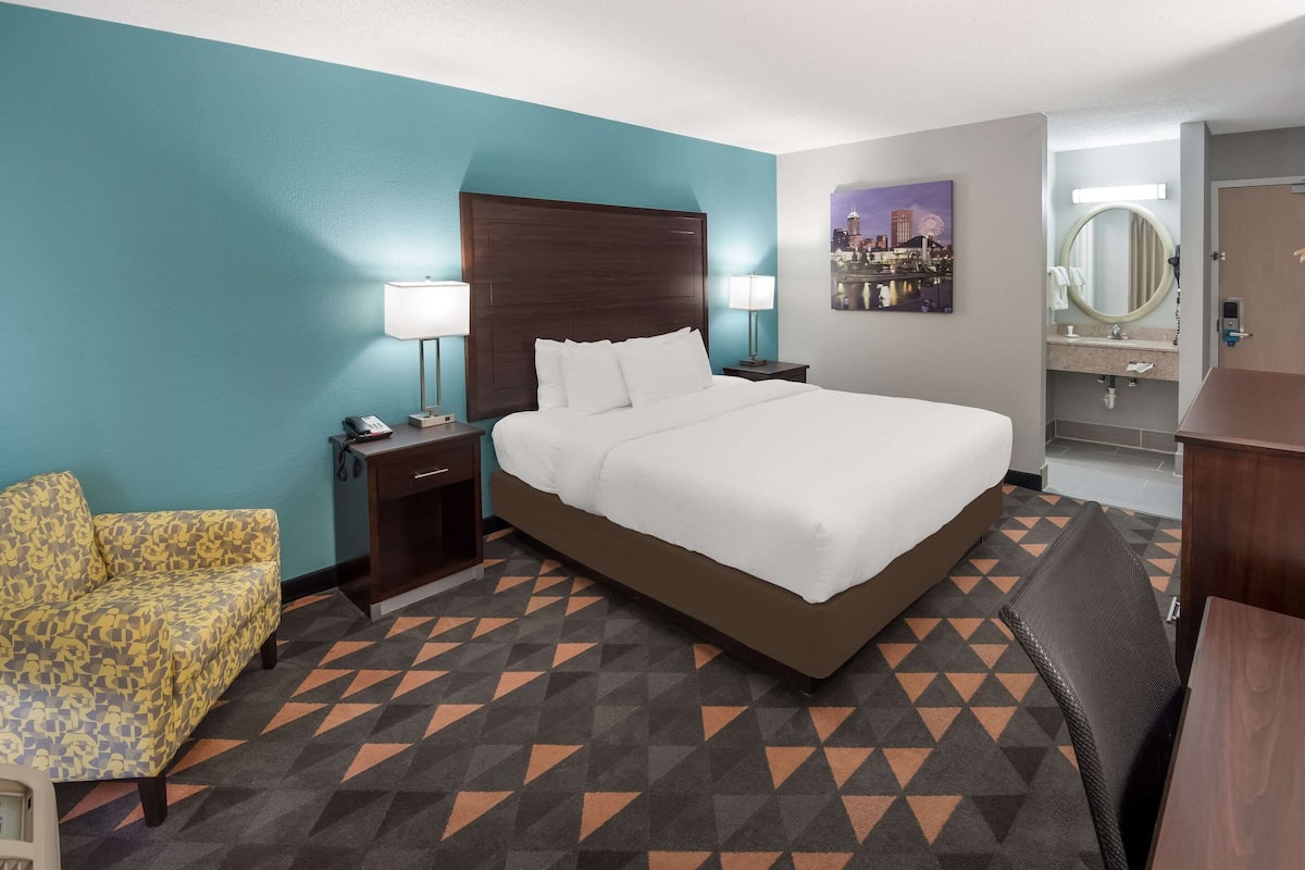 A hotel room with a large bed, a nightstand with a lamp, a chair, a bathroom with a mirror, and a cityscape painting on the wall.