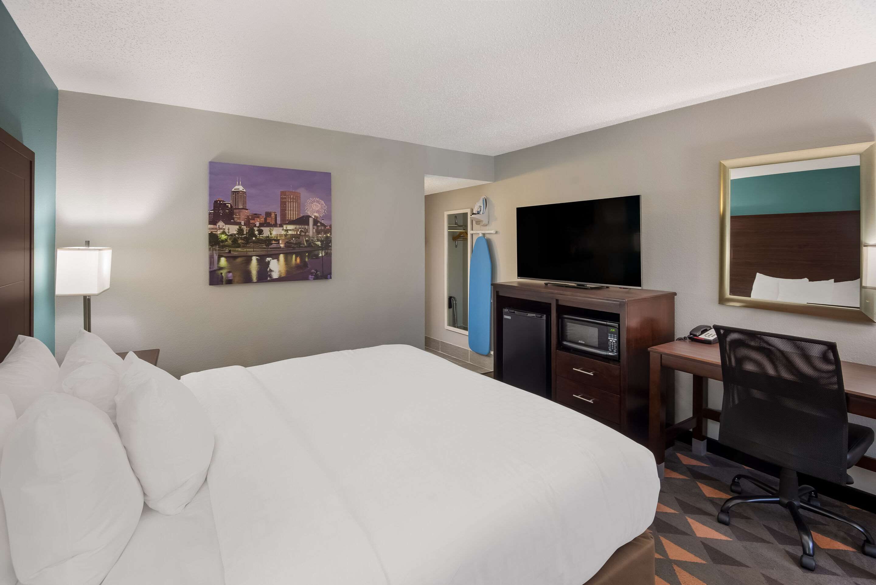 A hotel room with a bed, a desk, a television, a microwave, and a cityscape painting on the wall.