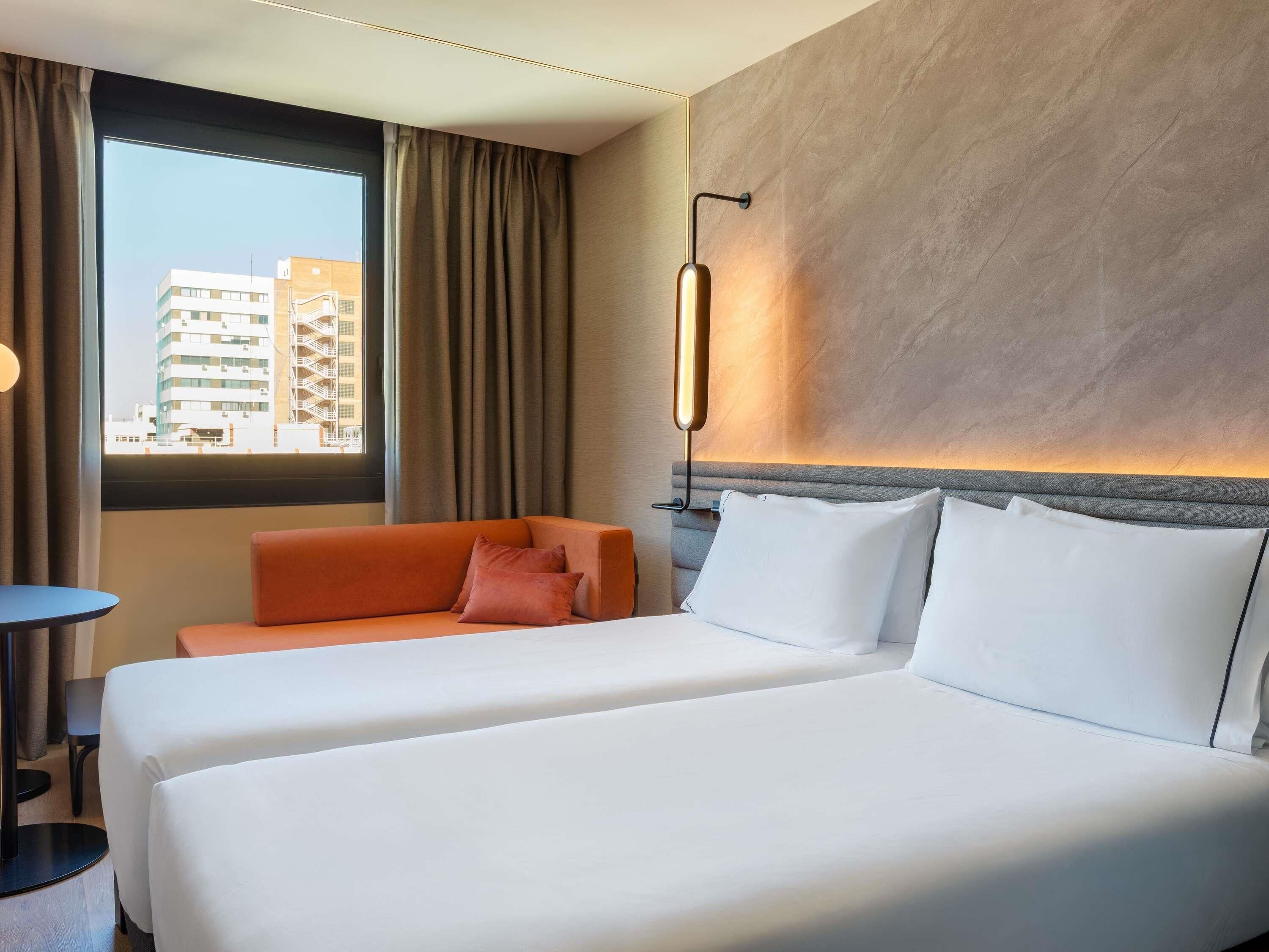 Habitació d'hotel amb un llit gran, un sofà, una taula petita i vista a edificis a través de la finestra.
