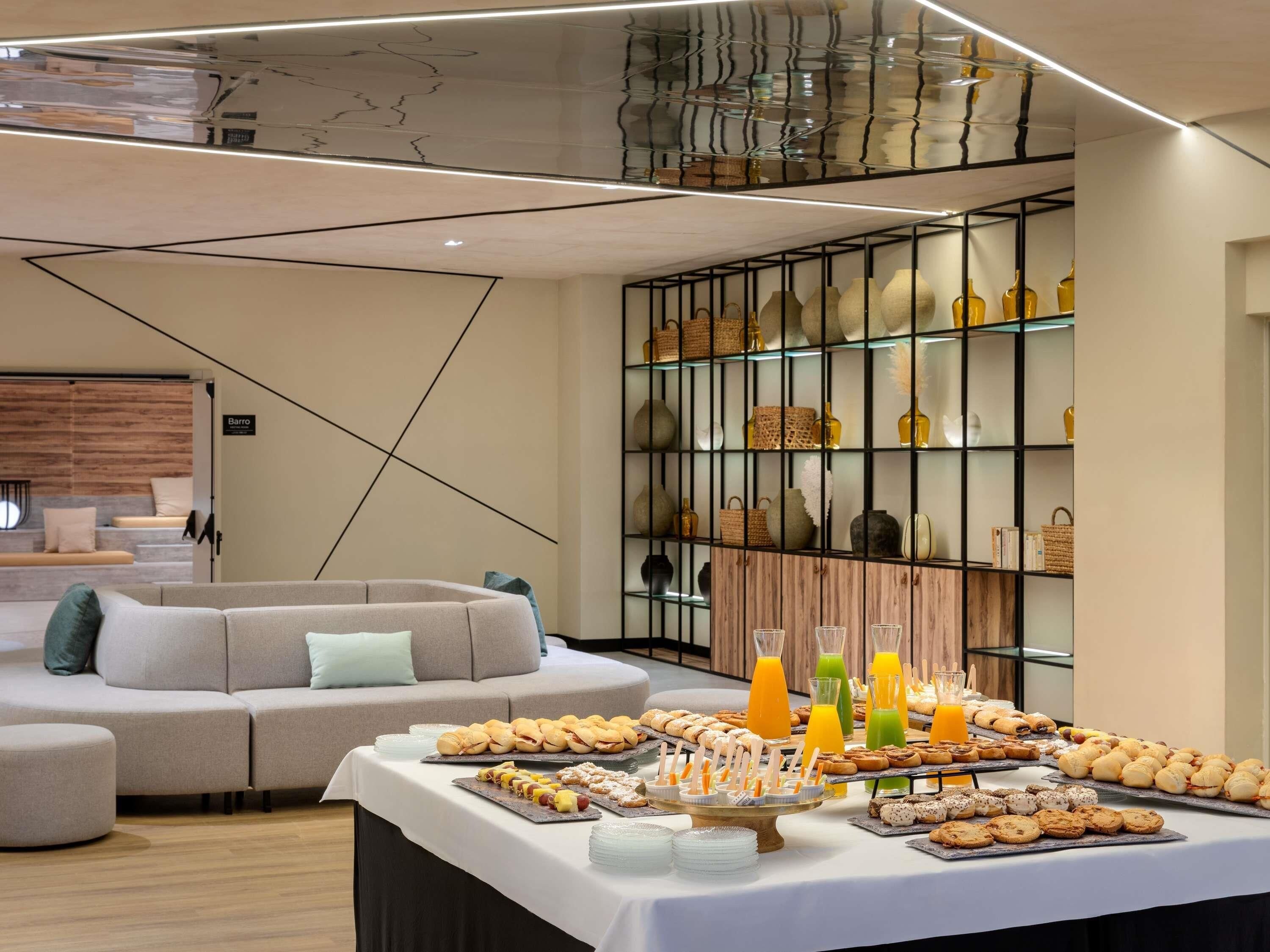 Una habitación de hotel moderna con un sofá, una mesa de buffet con pasteles y bebidas, y una pared decorativa con estanterías.