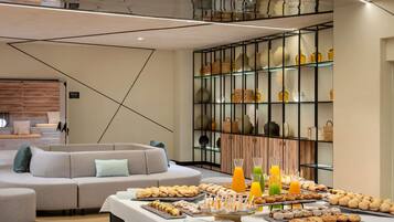 Una habitación de hotel moderna con un sofá, una mesa de buffet con pasteles y bebidas, y una pared decorativa con estanterías.