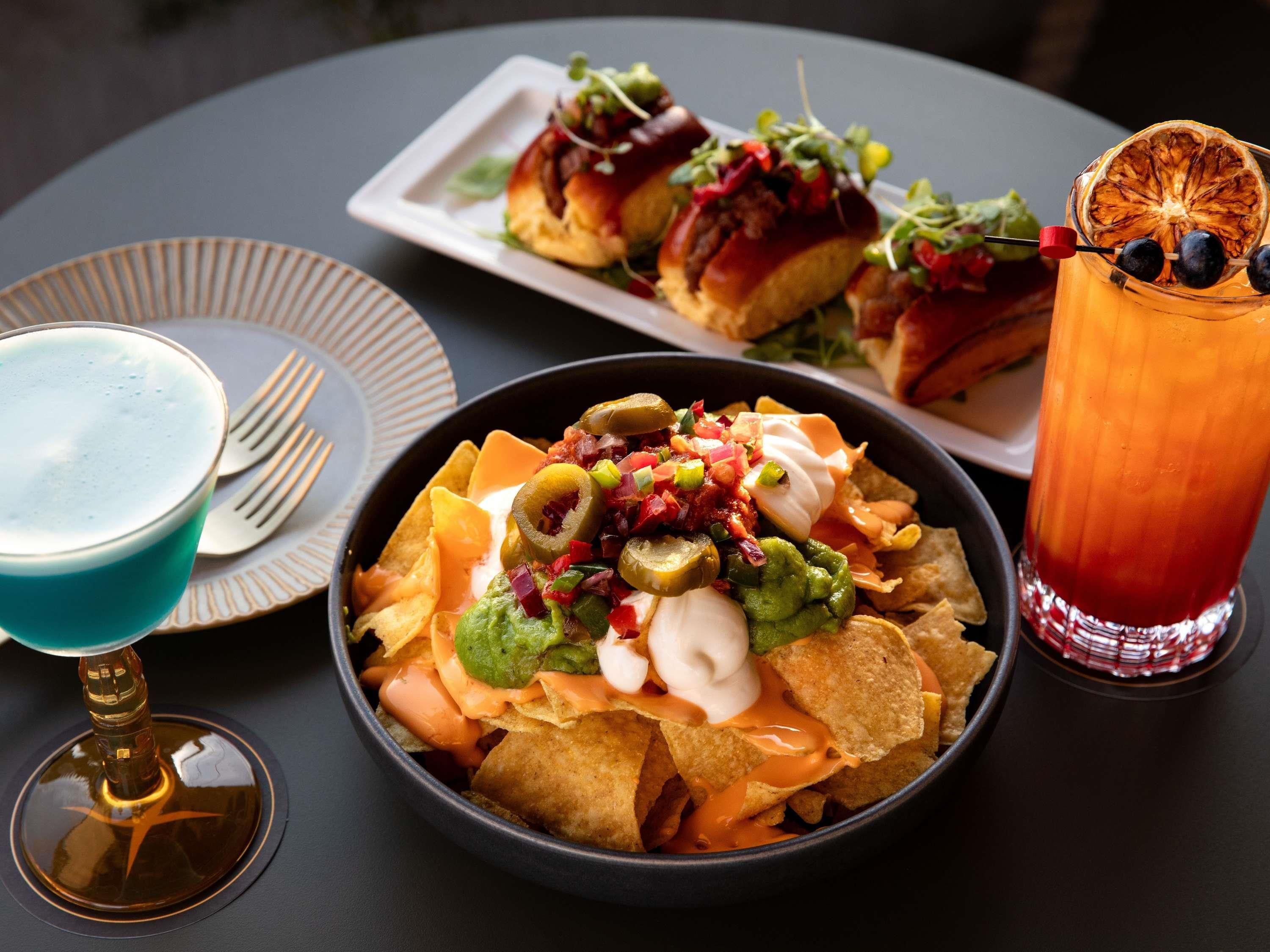 A table with nachos, a blue drink, a red drink, and a plate of food.