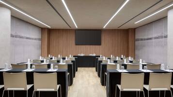 Un salón de conferencias con mesas, sillas, una pantalla de proyector y una pared de madera.