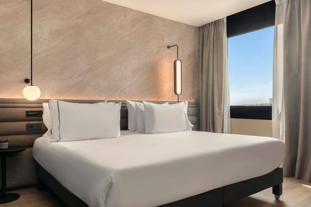 Une chambre d’hôtel moderne avec un grand lit, une table de chevet, une lampe fixée au mur et une fenêtre avec des rideaux.