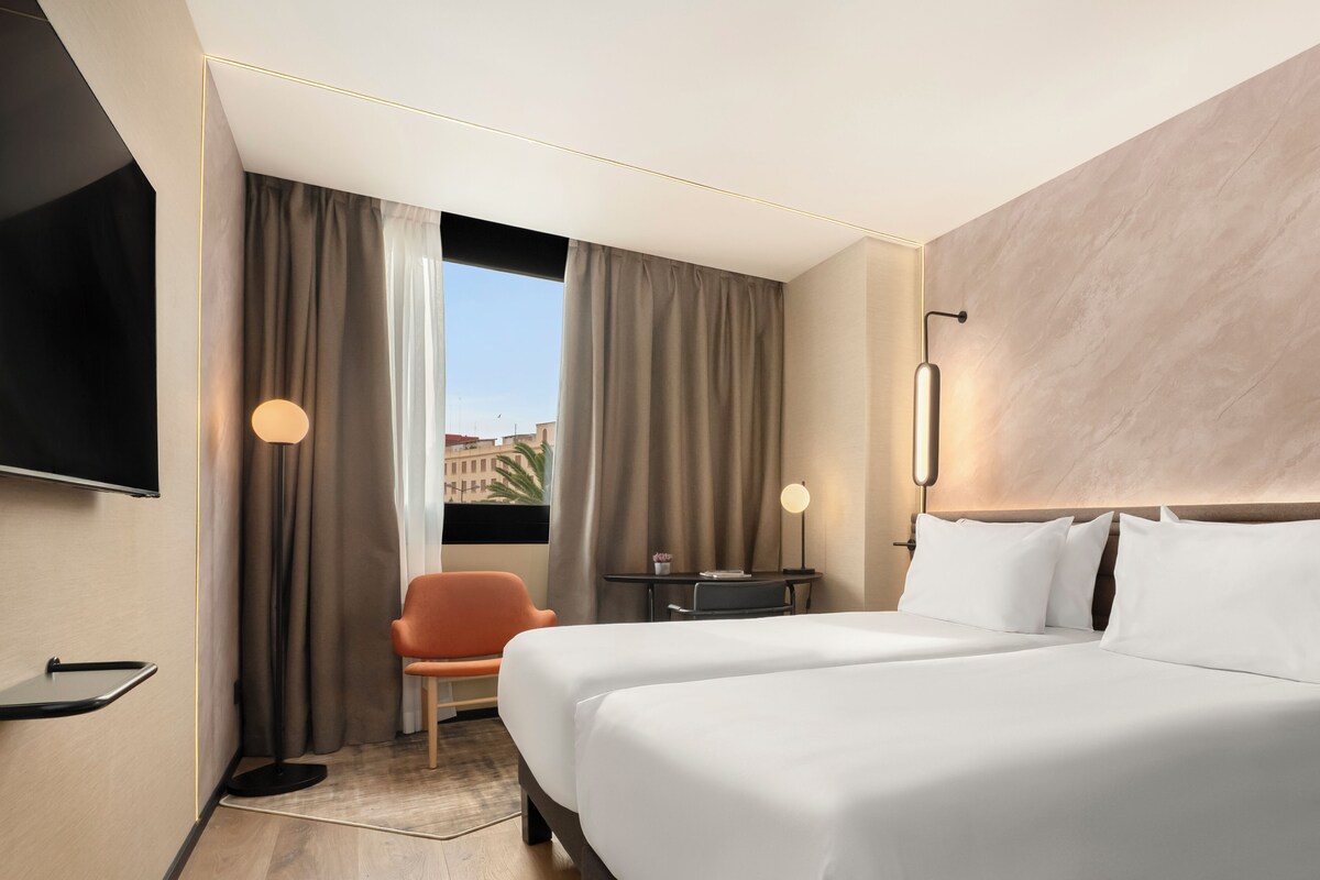 Habitación de hotel con dos camas, televisión, escritorio, silla y vista a la ciudad.