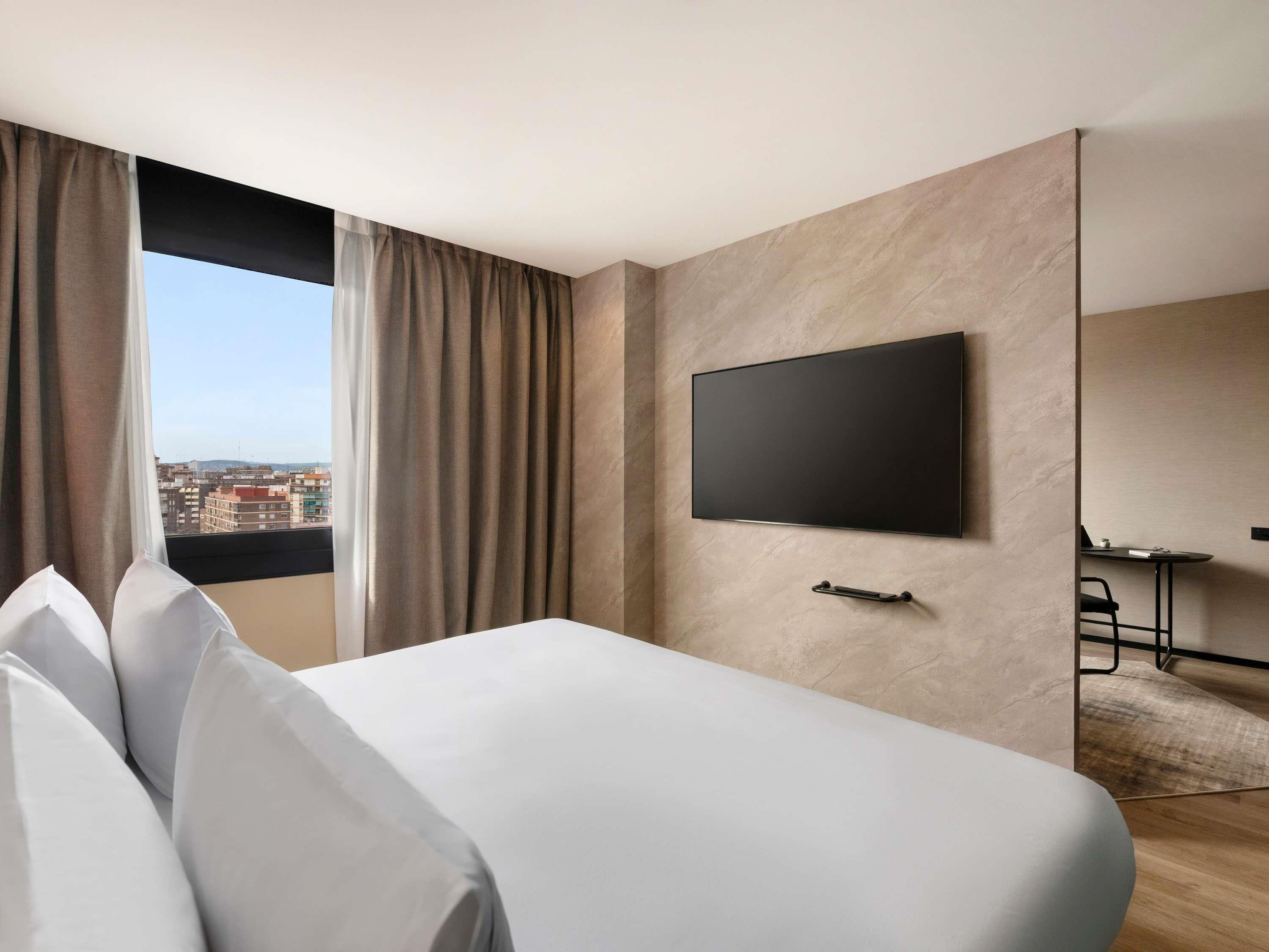 Habitación de hotel con cama, televisión en la pared, escritorio con silla y vista a la ciudad por la ventana.